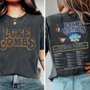 May include: A charcoal gray t-shirt featuring the text "LUKE COMBS" in a brown, vintage-style font on the front. The back of the shirt displays a concert graphic with the text "MY KINDA SATURDAY NIGHT" and a list of tour dates.