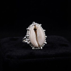 Può includere: Un anello in argento con una conchiglia di ciprea centrale. La conchiglia è di forma ovale con una superficie color crema e una fessura centrale marrone scuro. L'incastonatura in argento è decorata con piccole perline metalliche.