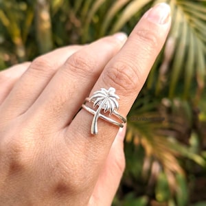 Puede incluir: Un anillo de plata con un diseño de palmera. El anillo tiene una doble banda y la palmera es el punto focal. El anillo se lleva en un dedo, con un fondo verde borroso.