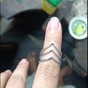 May include: A silver ring featuring a chevron design. The ring has three stacked chevron layers, creating a layered appearance. The ring is worn on a finger, highlighting its design and craftsmanship. The ring is made of a shiny, metallic material.