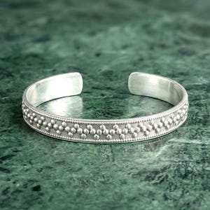 Puede incluir: Una pulsera tipo brazalete de plata con una banda texturizada que presenta un patrón repetitivo de pequeños puntos en relieve dispuestos en zigzag a lo largo del centro. La pulsera tiene un acabado pulido en el interior y extremos redondeados.