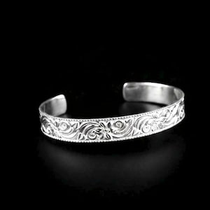 Könnte beinhalten: Ein silberfarbenes Armband in Manschettenform mit einem detaillierten, geprägten Blumen- und Rankenmuster auf der Außenseite. Das Metallband hat eine polierte Oberfläche, eine gleichmäßige Breite und eine offene Rückseite für das Handgelenk.