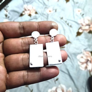 May include: A pair of silver rectangular earrings with a small round stud at the bottom of each rectangle. The earrings have a small round stud at the top that connects to the ear wire. The earrings are displayed against a floral background.