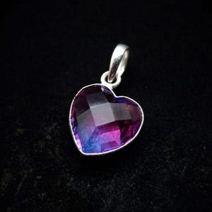 May include: A heart-shaped pendant featuring a faceted gemstone with a gradient color transition from deep purple to vibrant blue. The stone is set in a polished silver-toned metal frame with a small integrated bail at the top for attachment.
