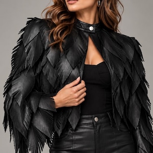 May include: A black jacket featuring a layered, feather-like fringe design throughout the sleeves and body. The jacket has a high collar with a single silver-toned snap button closure at the neck and an open front, worn over a black top and black trousers.