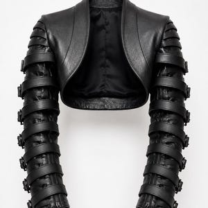 May include: A black leather bolero jacket featuring long sleeves adorned with multiple horizontal buckled straps. The garment has a smooth, open front design with a rounded neckline and a satin-like interior lining, showcasing a structured silhouette.