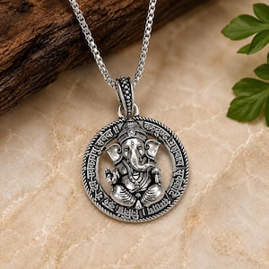 May include: A silver-toned pendant necklace featuring a circular frame with embossed Sanskrit text surrounding a central figure of Ganesha. The pendant hangs from a silver box chain, showcasing an oxidised finish that highlights the intricate details.