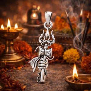 Puede incluir: Un colgante plateado con un diseño detallado de un tridente, un tambor y una mano sosteniendo un bastón. El colgante está intrincadamente elaborado con detalles ornamentales y un lazo para una cadena. El fondo incluye velas, flores e incienso.