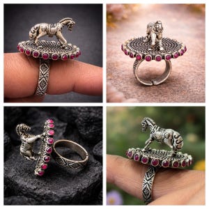 May include: A silver-toned ring featuring a horse figurine atop a circular platform adorned with red gemstones. The ring's band has an intricate, patterned design. The horse is detailed, and the gemstones are evenly spaced around the platform.