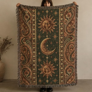 May include: A rectangular woven throw blanket featuring a dark green central panel with a sun and crescent moon design surrounded by stars. The side panels display a floral paisley pattern in shades of orange, brown, and green with fringe.