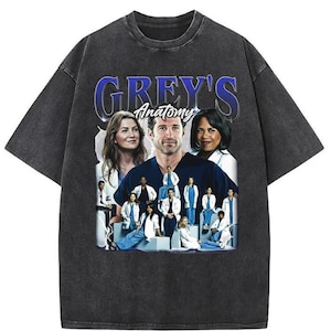 May include: Dark gray t-shirt featuring a graphic of the cast from the television show "Grey's Anatomy." The design includes the show's title in blue and images of the actors. The shirt has a vintage, washed look.