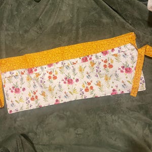 May include: A floral print apron with a white base and colorful flowers, including pink, orange, and purple. The apron has a yellow floral patterned top and ties. The apron is designed for kitchen use.