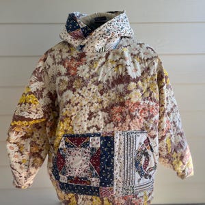 Könnte beinhalten: Ein gesteppter Kapuzenpullover mit einem Blumenmuster in Creme-, Braun-, Gelb- und Rottönen. Der Hoodie hat eine Patchwork-Tasche und einen hohen Kragen. Der Stoff wirkt weich und bequem, geeignet für Freizeitkleidung.
