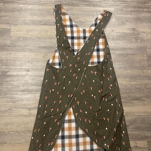 May include: Reversible apron with a green mushroom print on one side and a plaid pattern on the other. The apron has a cross-back design and is made of fabric. The color palette includes green, brown, white, and orange.