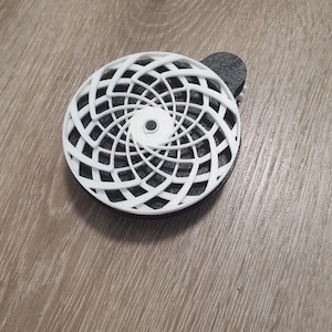 Può includere: Un design geometrico bianco, stampato in 3D, è posto su una base circolare nera. Il design presenta un motivo a spirale di linee interconnesse. Un piccolo pezzo semicircolare nero è attaccato al lato. Su una superficie di legno.