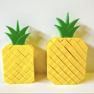 May include: Two yellow pineapple-shaped ornaments with green leafy tops. The pineapples are made from small, square blocks, creating a textured surface. One is rectangular, the other is trapezoidal.
