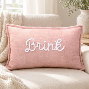 May include: A rectangular throw pillow with a textured dusty pink fabric cover. The center of the pillow features the word "Brink" embroidered in white cursive script lettering. The pillow has a piped edge finish and a consistent woven fabric texture.