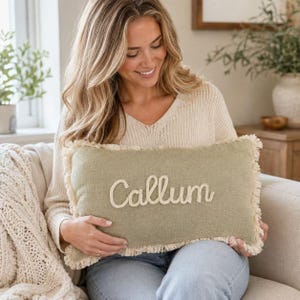 May include: A rectangular sage green throw pillow featuring a frayed edge trim and the name Callum embroidered in a cream-coloured script font across the centre. The pillow has a textured fabric surface and a soft, woven appearance.