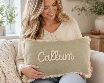 May include: A rectangular sage green throw pillow featuring a frayed edge trim and the name Callum embroidered in a cream-colored script font across the center. The pillow has a textured fabric surface and a soft, woven appearance.
