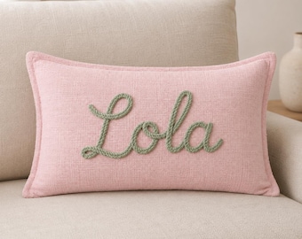 May include: A rectangular light pink throw pillow with a textured fabric finish. The name Lola is embroidered in the center using a thick, sage green rope-like yarn in a cursive script font, creating a raised and tactile decorative effect.