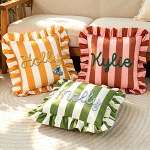 May include: Three square throw pillows with ruffled edges and vertical stripes. One is yellow and white with the name Holly and blue flower accents. One is brown and pink with the name Kylie. One is green and white with the name molly.