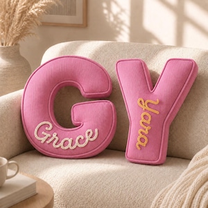 May include: Two pink corduroy letter-shaped pillows, one in the shape of the letter G with the name Grace embroidered in white, and one in the shape of the letter Y with the name Yara embroidered in yellow, both featuring a soft, ribbed texture.