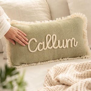 May include: A rectangular sage green throw pillow featuring a textured fabric surface and cream-colored fringe trim along the edges. The name Callum is embroidered in a raised, cream-colored cursive rope-style script across the center front.
