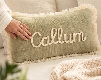 May include: A rectangular sage green throw pillow featuring a textured fabric surface and cream-colored fringe trim along the edges. The name Callum is embroidered in a raised, cream-colored cursive rope-style script across the center front.