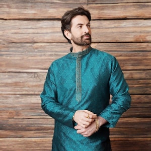 Può includere: Kurta color teal con motivo floreale. La tunica a maniche lunghe presenta un colletto alla coreana e ricami dorati lungo lo scollo e la paramontura. La kurta è indossata con bracciali e un anello dorati.