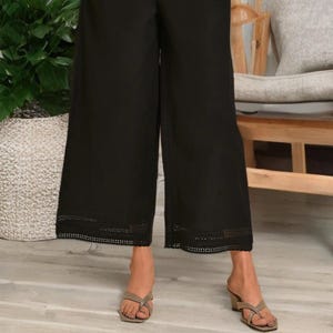 May include: Black wide-leg pants with a decorative, perforated hemline. The pants have pockets and are worn with neutral-toned sandals. The model is wearing a yellow top. The pants are a stylish addition to any wardrobe.