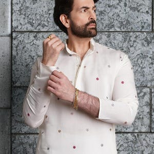 Pode incluir: Kurta de manga comprida creme com pequenos padrões florais bordados em vermelho, dourado e cinza. A kurta apresenta uma gola mandarim e uma carcela abotoada. Uma pulseira e um anel dourados são visíveis.