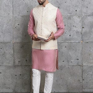 May include: A formal ensemble featuring a cream-colored vest with a geometric pattern over a pink long-sleeved shirt and matching pink kurta. The outfit is completed with white pants and black sandals. The man is holding a book.