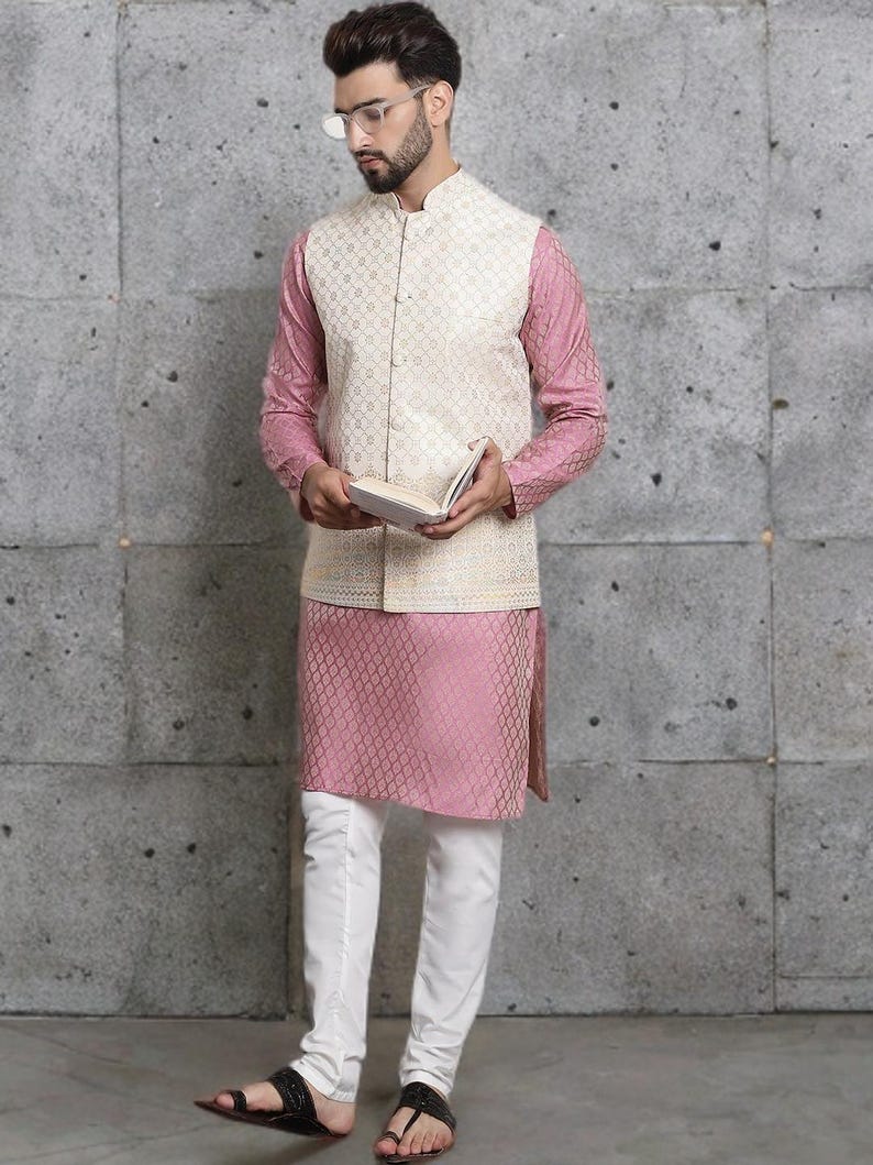 May include: A formal ensemble featuring a cream-colored vest with a geometric pattern over a pink long-sleeved shirt and matching pink kurta. The outfit is completed with white pants and black sandals. The man is holding a book.