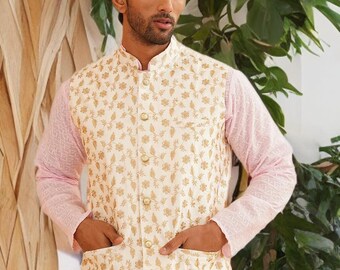 May include: A cream-colored vest with gold floral embroidery, worn over a light pink long-sleeved shirt. The vest has a mandarin collar and two front pockets. The outfit is part of a traditional South Asian ensemble.