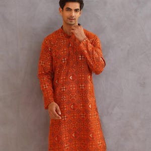 May include: An orange kurta with a geometric pattern. The long-sleeved shirt has a mandarin collar and is paired with white pants. The kurta features a repeating design in black and gold.
