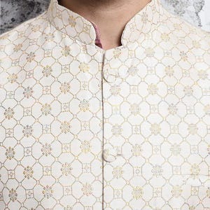 May include: Close-up of a cream-colored, button-down jacket with an intricate floral and geometric pattern in gold and other muted colors. The jacket has a mandarin collar and a pink lining visible at the neckline.