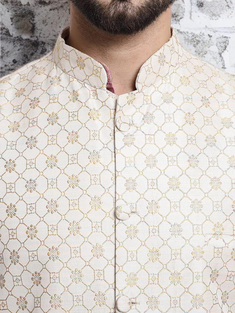 May include: Close-up of a cream-colored, button-down jacket with an intricate floral and geometric pattern in gold and other muted colors. The jacket has a mandarin collar and a pink lining visible at the neckline.