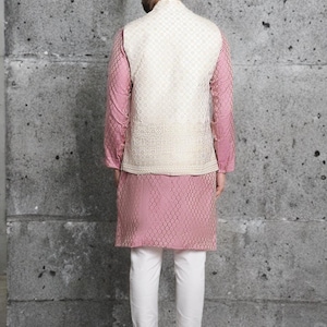 May include: A formal ensemble featuring a pink kurta and matching long-sleeved shirt, paired with white pants. A cream-colored, intricately patterned vest completes the outfit. The clothing is designed for a formal occasion.