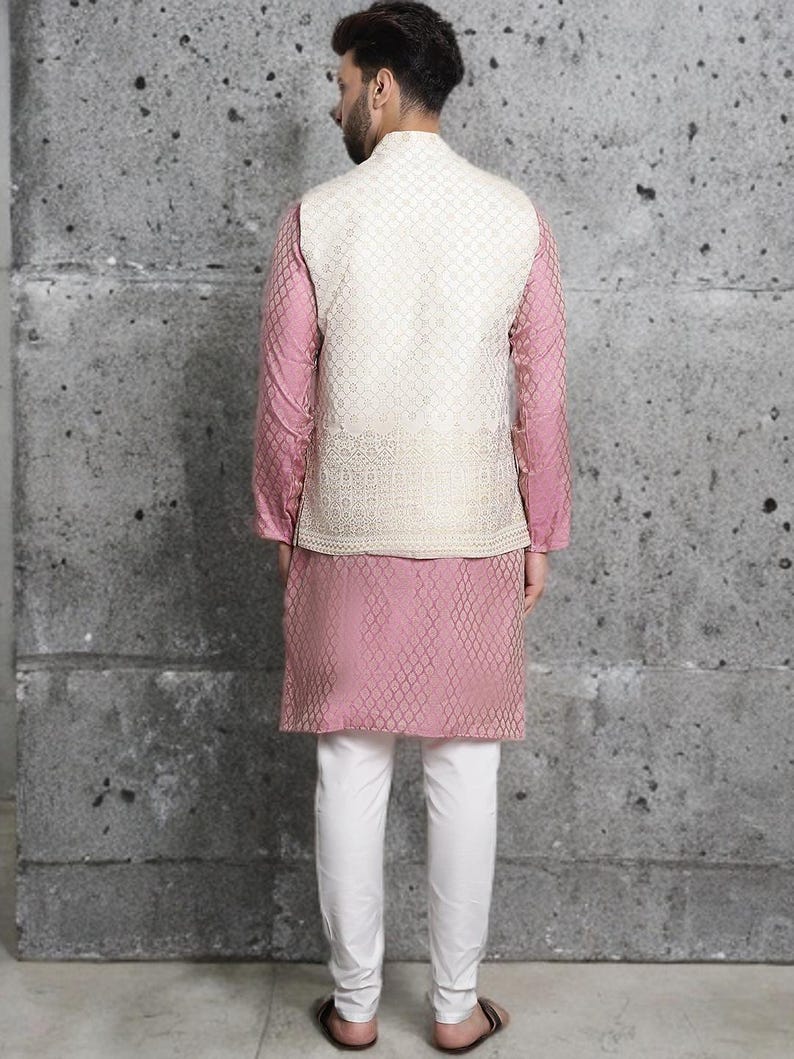 May include: A formal ensemble featuring a pink kurta and matching long-sleeved shirt, paired with white pants. A cream-colored, intricately patterned vest completes the outfit. The clothing is designed for a formal occasion.