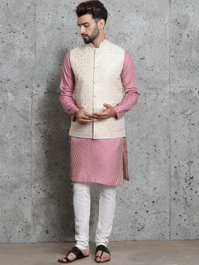 May include: A three-piece ensemble featuring a cream-colored vest with a geometric pattern over a pink long-sleeved shirt and matching pink tunic. The outfit is completed with white pants and black sandals. The background is a textured gray wall.