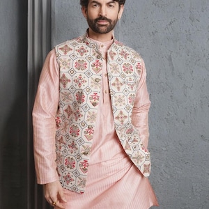 May include: A light pink kurta pajama set with a patterned vest. The vest has a cream background and a repeating floral design in red, pink, and gold. The kurta has a mandarin collar and long sleeves. The outfit is suitable for formal events.