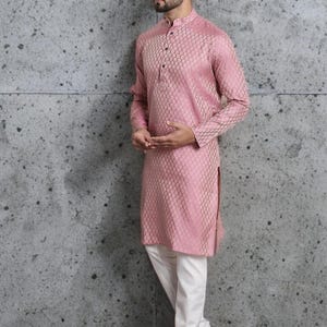 May include: A pink long-sleeved kurta with a geometric pattern, paired with white pants. The kurta has a mandarin collar and buttoned placket. The outfit is completed with brown sandals. The background is a textured gray wall.
