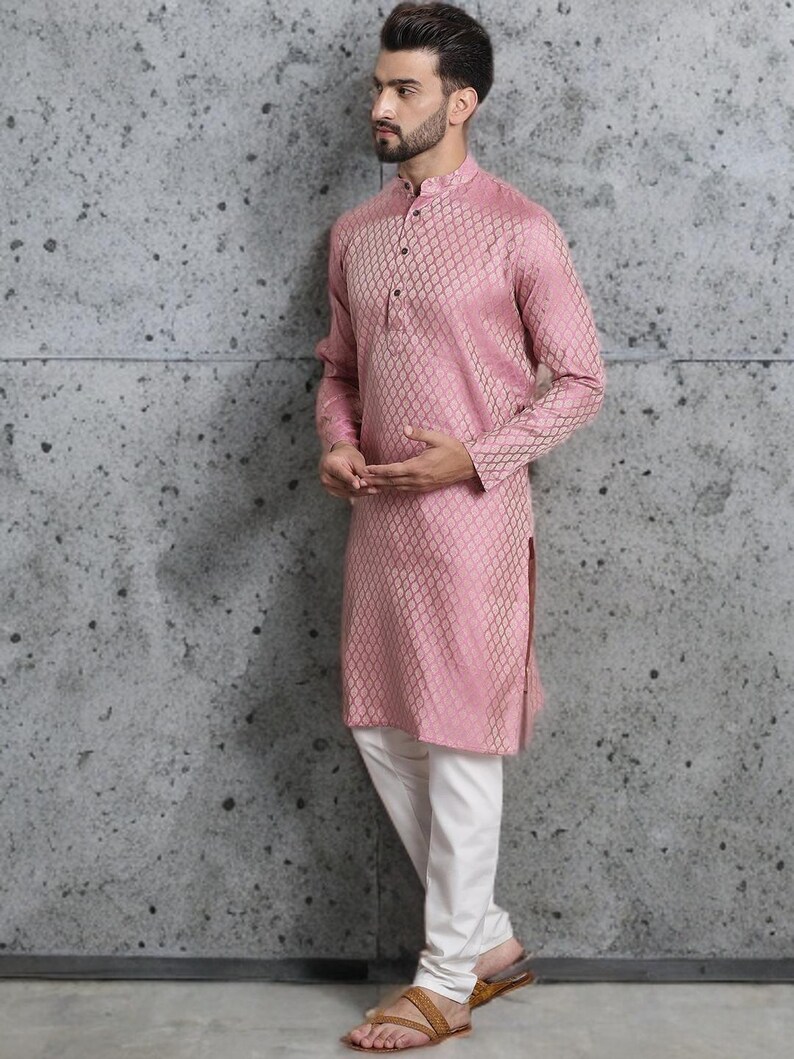 May include: A pink long-sleeved kurta with a geometric pattern, paired with white pants. The kurta has a mandarin collar and buttoned placket. The outfit is completed with brown sandals. The background is a textured gray wall.