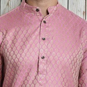 May include: A pink kurta with a gold-colored, ornate pattern. The long-sleeved shirt has a mandarin collar and three decorative buttons down the front. The fabric has a subtle sheen.