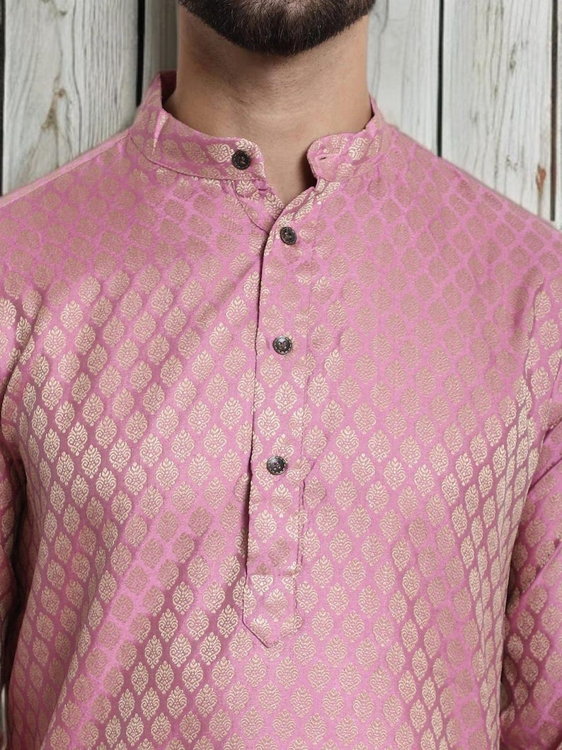 May include: A pink kurta with a gold-colored, ornate pattern. The long-sleeved shirt has a mandarin collar and three decorative buttons down the front. The fabric has a subtle sheen.