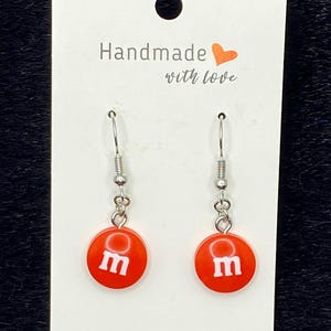 May include: A pair of handmade earrings with red circular charms featuring a white letter "m" design. The earrings hang from silver-colored fish hook ear wires. The backing card reads "Handmade with love."