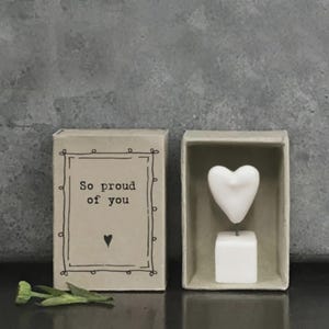 Könnte beinhalten: Eine kleine, offene Streichholzschachtel mit dem Text "So proud of you" und einem Herzsymbol. Daneben befindet sich ein weißes herzförmiges Objekt auf einem kleinen Ständer in einer ähnlichen Schachtel. Eine getrocknete Blume liegt unten.