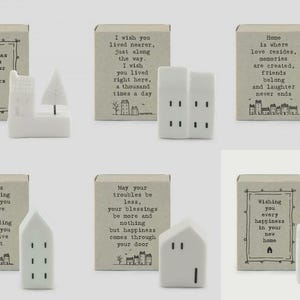 Puede incluir: Una colección de figuritas de casas de cerámica blanca y cajas de cerillas decorativas con citas inspiradoras. Las cajas de cerillas tienen textos como "Christmas wishes from our house to yours" y "Home is where love resides".