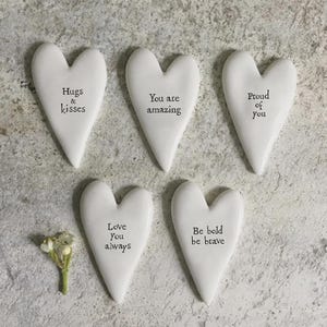 Könnte beinhalten: Fünf weiße herzförmige Dekorationsobjekte mit inspirierenden Botschaften in schwarzem Text. Die Botschaften lauten: "Hugs & kisses", "You are amazing", "Proud of you", "Love You always" und "Be bold be brave". Ein kleiner Zweig weißer Blumen ist ebenfalls vorhanden.