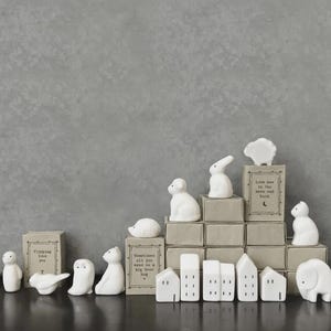 Puede incluir: Una colección de figuritas de cerámica blanca, que incluyen animales y casas en miniatura, dispuestas sobre una superficie oscura contra un fondo gris. Algunas figuritas están colocadas sobre pequeñas cajas con los textos "Flipping love you", "Sometimes all you need is a big bear hug" y "Love ave to the moon and back".